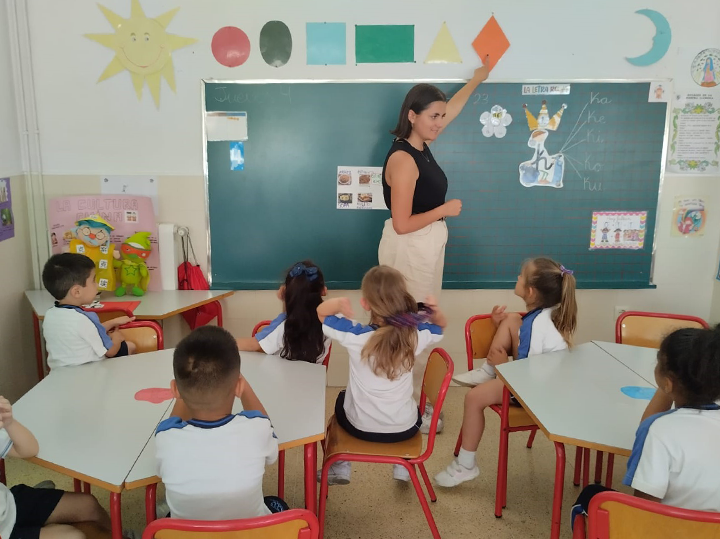 A student standing at the front of a classroom with children sitting in chairs in front of them.