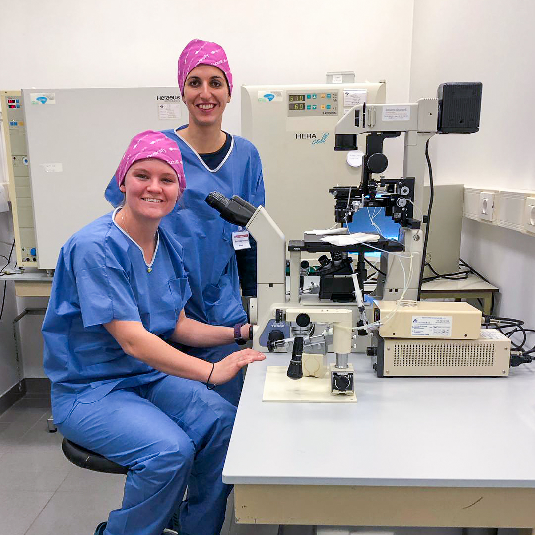 Two students in blue scrubs and pink bandana's on their heads in front of a microscope in a lab.