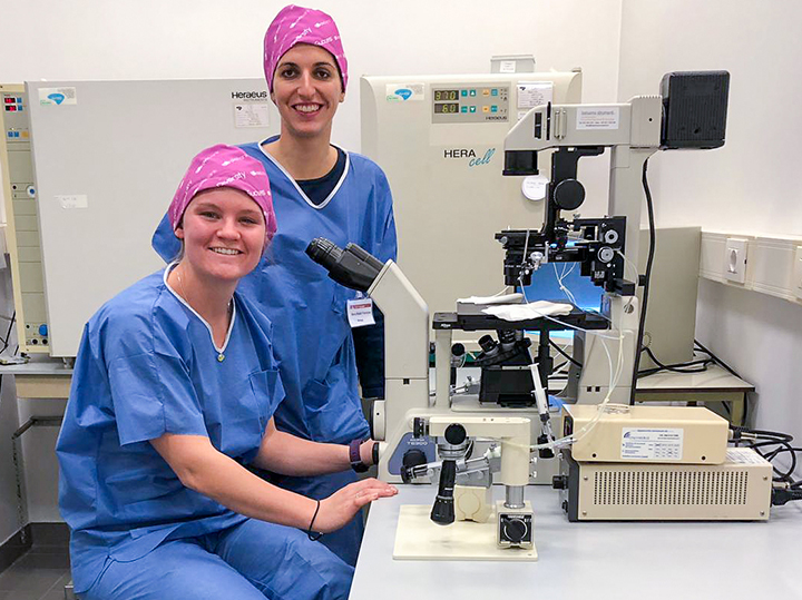 Two students in blue scrubs and pink bandana's on their heads in front of a microscope in a lab.