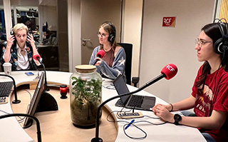 Three students sitting in front of microphones wearing headphones producing a radio segment in Lyon, France.