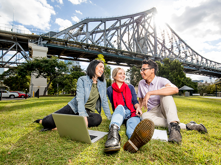 Three students sitting on the grass with a bridge behind them in Brisbane, Australia.