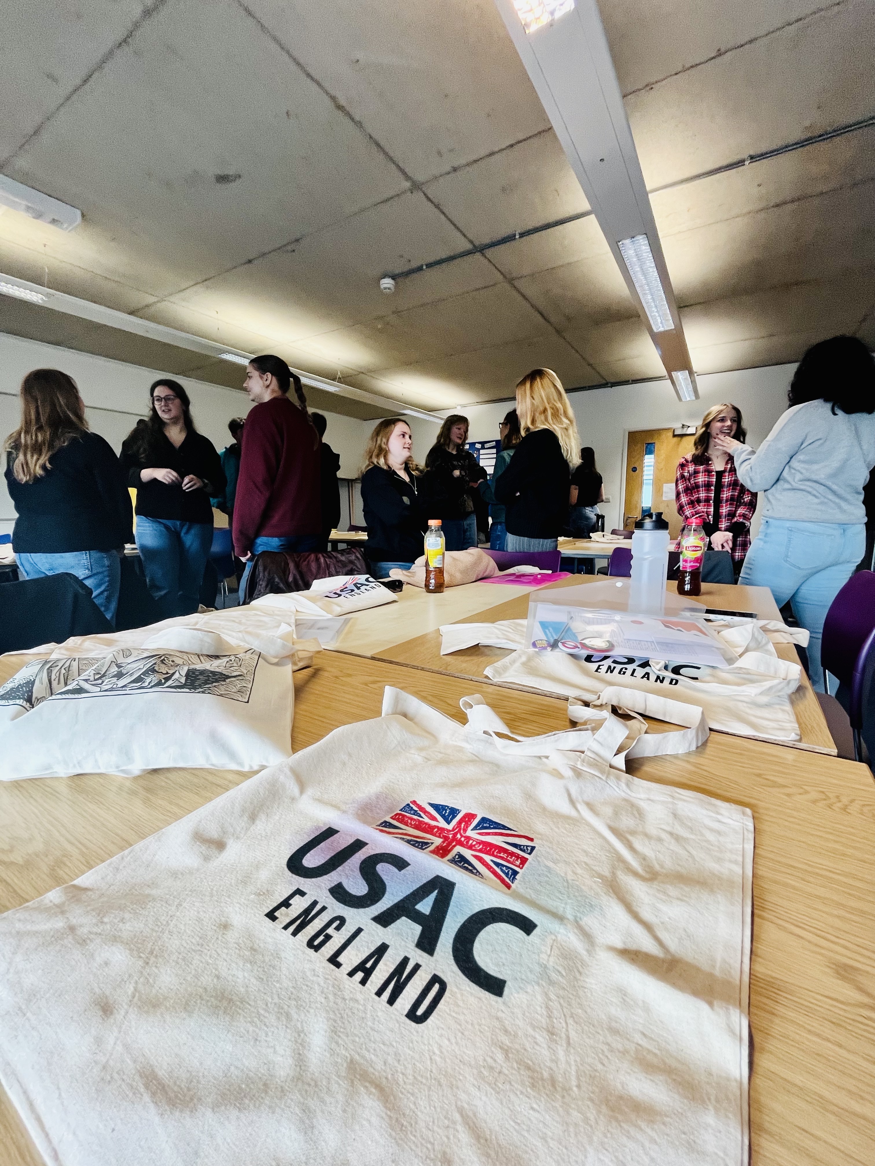 A table with a tote bag with USAC England and the British flag on it with students conversing in the background