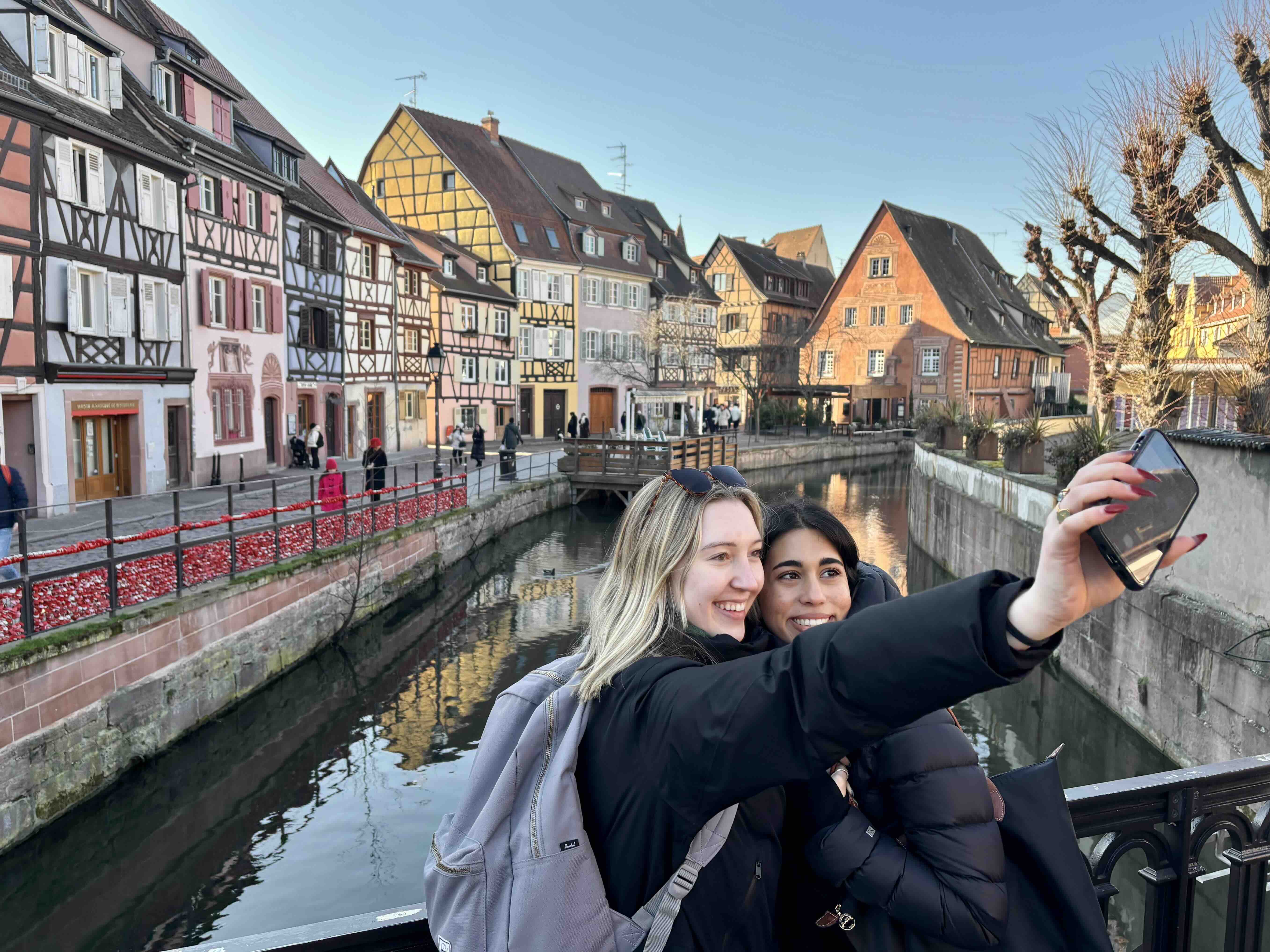 Two students taking a selfie with a view of traditional German-style buildings along a canal in Strasbourg, France.