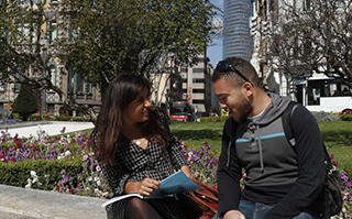 Two people sitting and conversing outside with city buildings in the background in Bilbao, Spain.