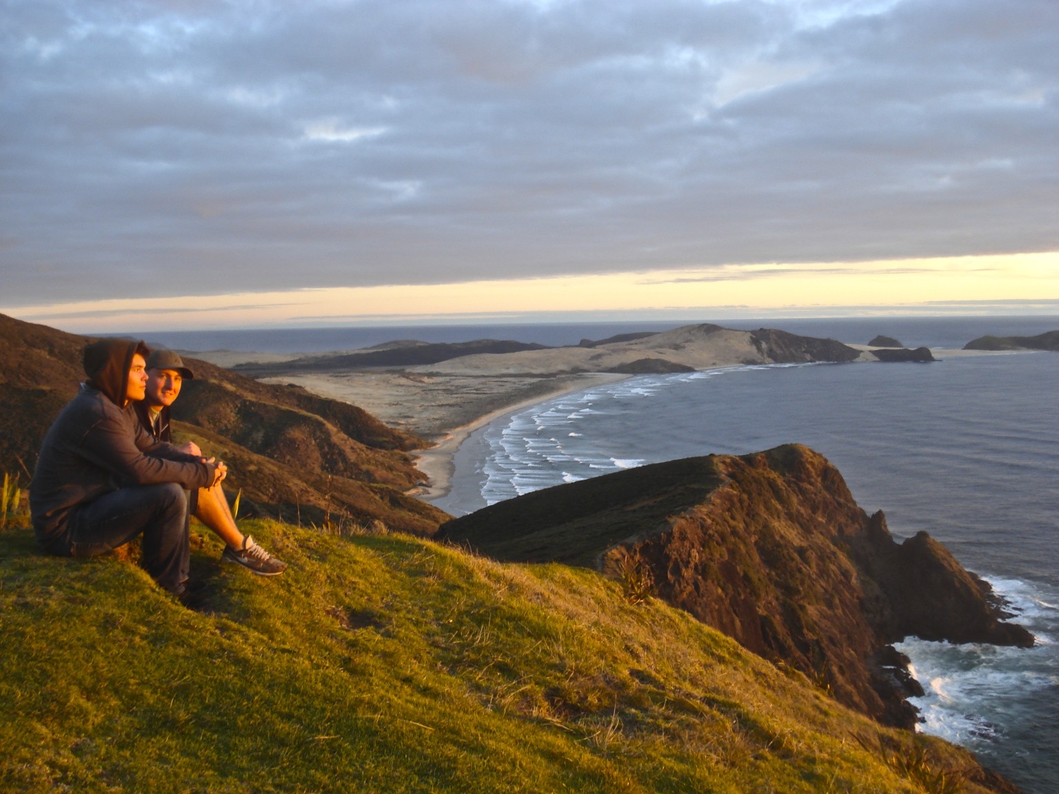 Two people sitting on a coastal cliff at sunset in Auckland, New Zealand.